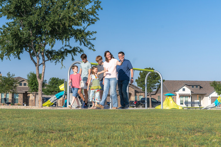 BC_02113-Enhanced-NR A family walks together through a neighborhood park on a sunny day, with children playing near colorful playground equipment and homes visible in the background.
