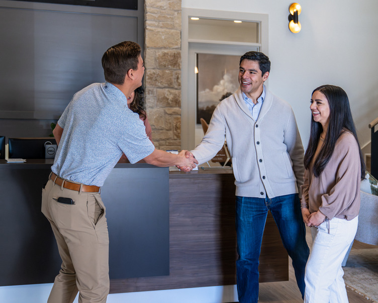 DSC01198-Enhanced-NR A homebuyer shakes hands with a sales representative inside a modern home sales office while another person smiles beside them.