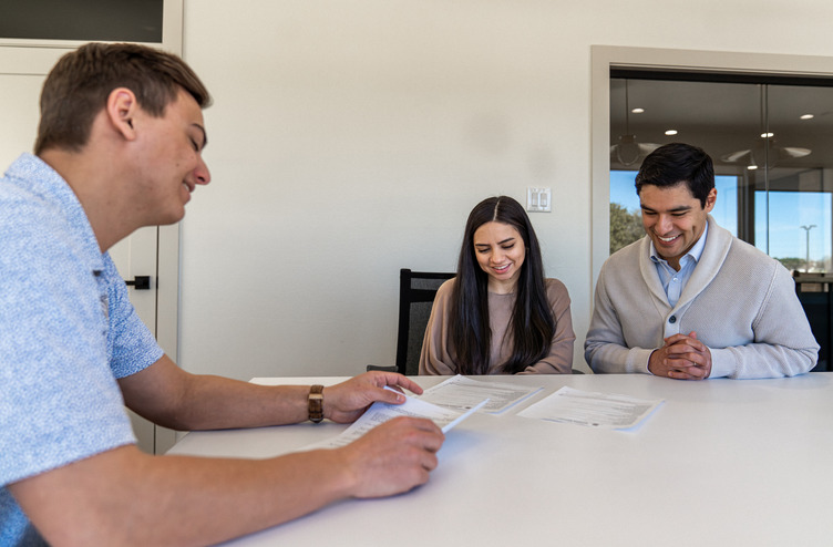 DSC01445-Enhanced-NR A Betenbough Homes team member reviews paperwork with a smiling couple seated at a table during a home buying appointment.